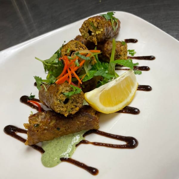 Indian seekh kebabs with green sauce, dark drizzle, fresh herbs, and a lemon wedge.