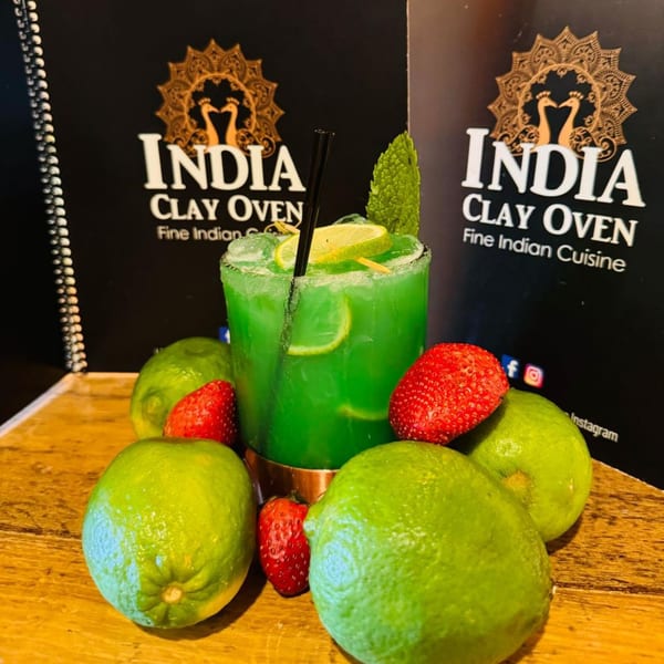 A green cocktail with lime slices and mint, surrounded by whole limes and strawberries, with India Clay Oven menus in the background.