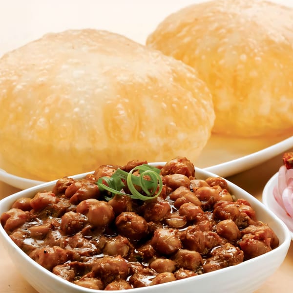 A white bowl of chana masala with green garnish, served with two fluffy puri breads.