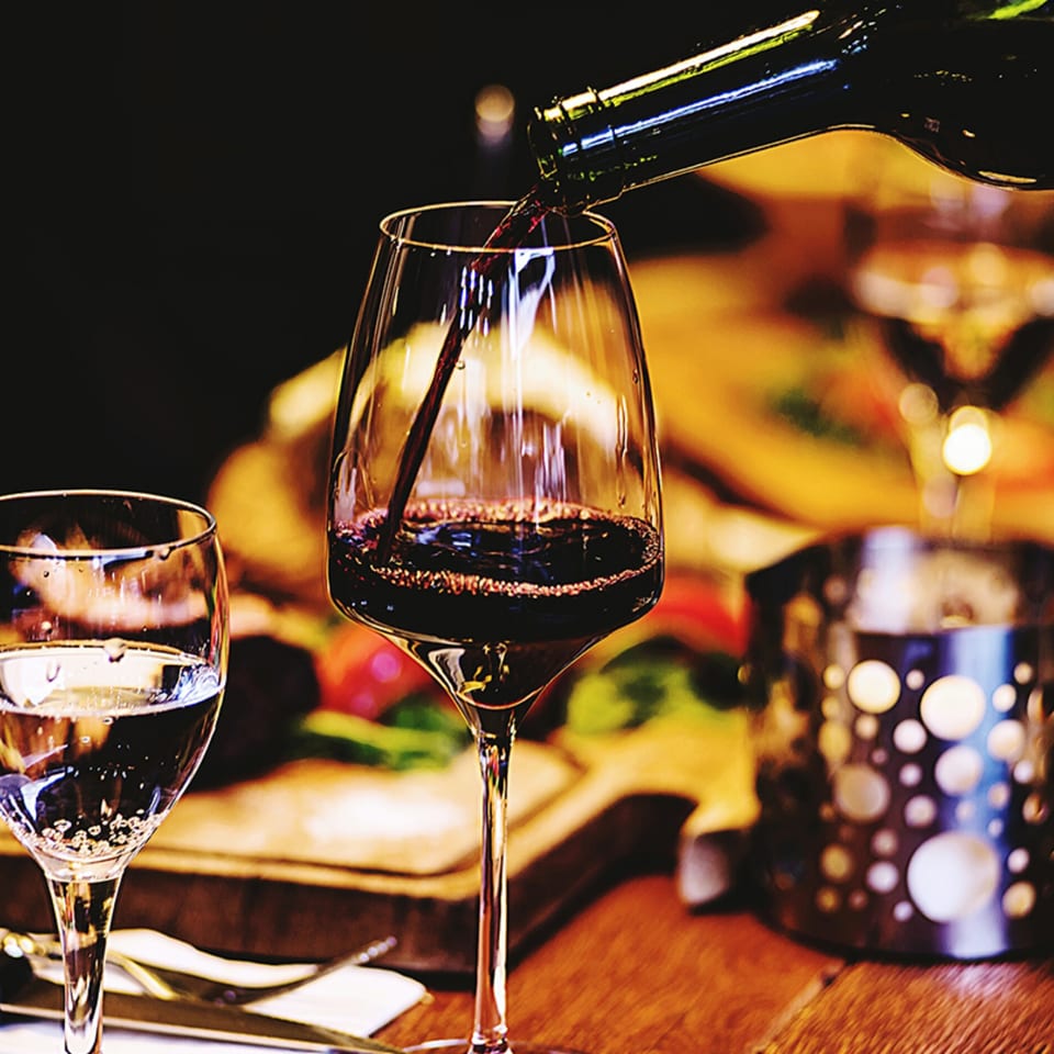Red wine being poured into a glass at a table with a water glass and candle holder.