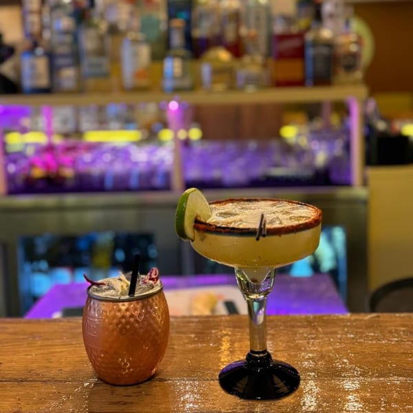 Two cocktails, a margarita and a copper mug drink, on a wooden bar with blurred bottles in background.